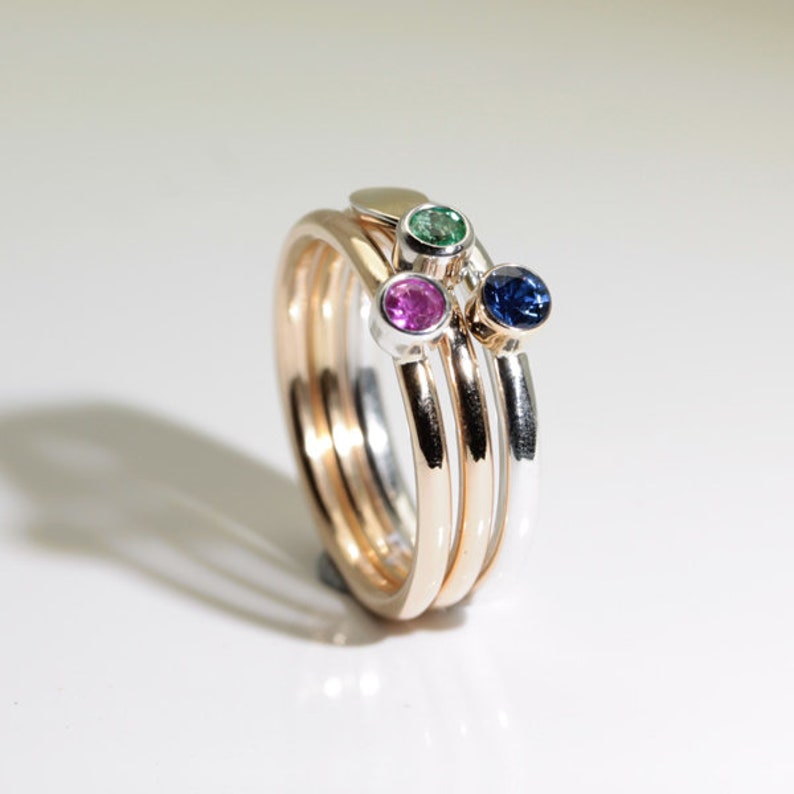 Może przedstawiać: Zestaw pierścionk&oacute;w do układania w stosy w odcieniach złota i srebra, każdy z kamieniem szlachetnym. Kamienie szlachetne są w kolorze granatowym, żywej zieleni i jasnor&oacute;żowym. Pierścionki są prezentowane na białej powierzchni.