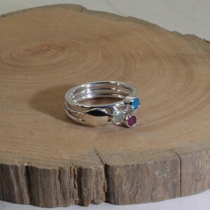 Może przedstawiać: Zestaw srebrnych pierścionk&oacute;w, kt&oacute;re można układać w stos. Każdy pierścionek ma inny kamień szlachetny: niebieski, biały i czerwony. Pierścionki są prezentowane na drewnianej powierzchni.