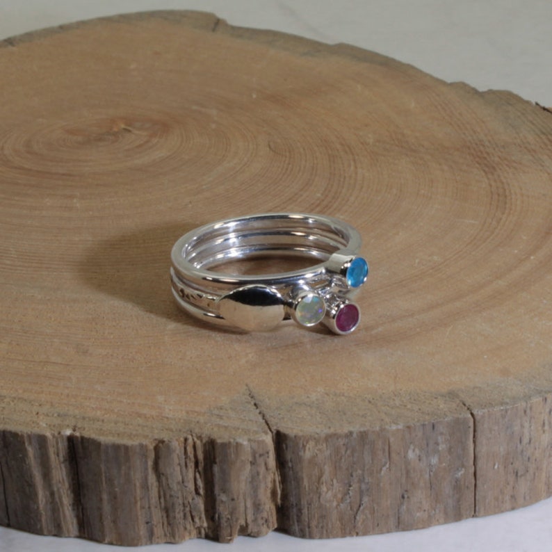 Może przedstawiać: Zestaw srebrnych pierścionk&oacute;w, kt&oacute;re można układać w stos. Każdy pierścionek ma inny kamień szlachetny: niebieski, biały i czerwony. Pierścionki są prezentowane na drewnianej powierzchni.