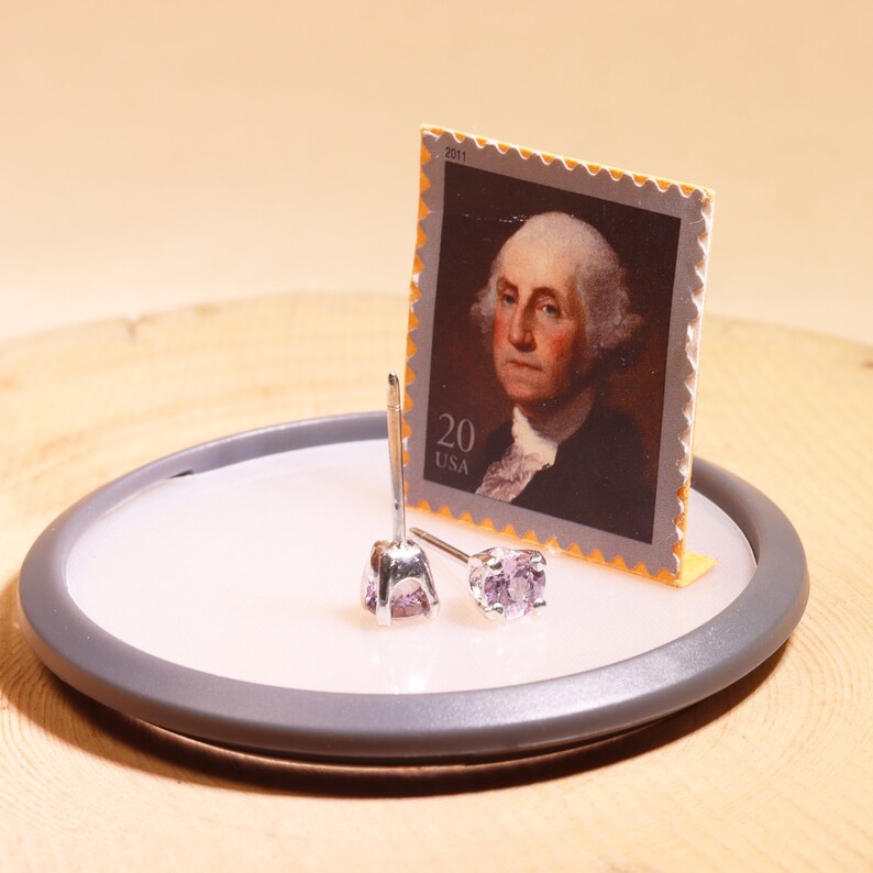 May include: A pair of silver stud earrings with small, light purple gemstones. The earrings are displayed on a small, round, gray tray. A 2011 US postage stamp featuring George Washington is in the background.