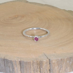 Może przedstawiać: Srebrny pierścionek z małym, okrągłym, czerwonym kamieniem szlachetnym. Pierścionek leży na jasnej drewnianej powierzchni. Opaska jest cienka, a kamień szlachetny jest centralnym punktem.