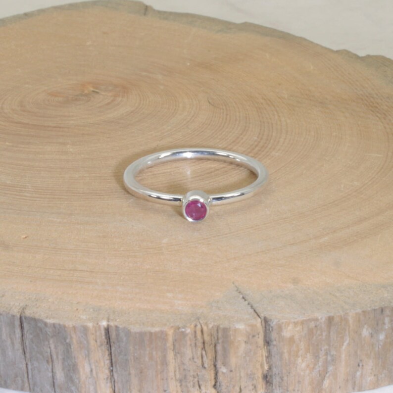 Może przedstawiać: Srebrny pierścionek z małym, okrągłym, czerwonym kamieniem szlachetnym. Pierścionek leży na jasnej drewnianej powierzchni. Opaska jest cienka, a kamień szlachetny jest centralnym punktem.