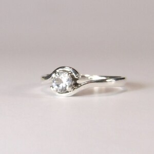 May include: A silver ring with a single, clear, round gemstone set in a curved band.