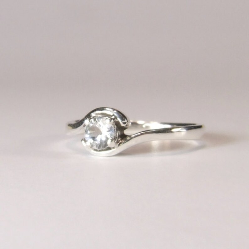May include: A silver ring with a single, clear, round gemstone set in a curved band.