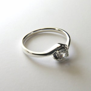May include: A silver ring with a single, clear, round gemstone set in a prong setting. The band is thin and has a curved design.