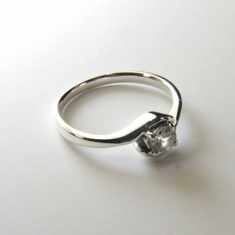 May include: A silver ring with a single, clear, round gemstone set in a prong setting. The band is thin and has a curved design.