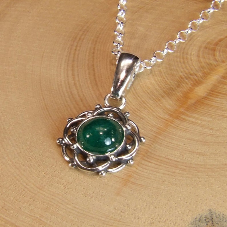 K&ouml;nnte beinhalten: Eine silberne Halskette mit einem gr&uuml;nen Edelstein-Anh&auml;nger. Der Anh&auml;nger ist ein runder Cabochon-Stein, der in einem filigranen Silberdesign gefasst ist.