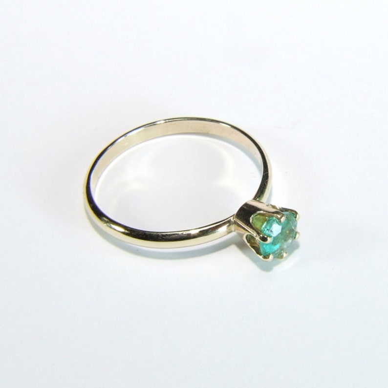 Pode incluir: Um anel de ouro com uma pedra preciosa redonda verde claro. O anel tem uma banda simples e uma crava&ccedil;&atilde;o de quatro garras que segura a pedra preciosa. O anel est&aacute; sobre um fundo branco.
