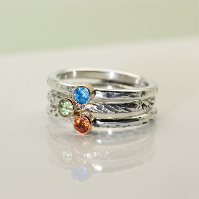 Może przedstawiać: Stos srebrnych pierścionk&oacute;w z teksturowanymi obrączkami. Każdy pierścionek ma mały, kolorowy kamień szlachetny: niebieski, zielony i pomarańczowy. Kamienie szlachetne są osadzone w oprawach w kolorze złota. Pierścionki są prezentowane na odblaskowej powierzchni.