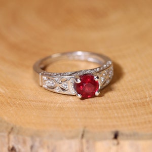 May include: A silver ring featuring a large, red gemstone surrounded by smaller, clear gemstones. The ring has a delicate, intricate design.