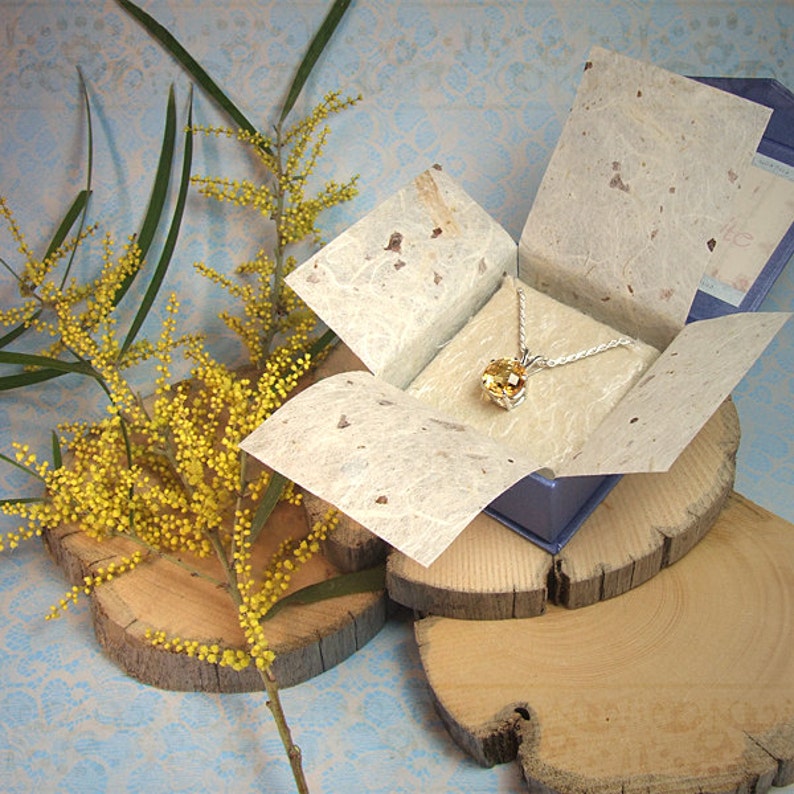K&ouml;nnte beinhalten: Eine goldene Halskette mit einem gro&szlig;en, runden, facettierten Edelstein in einer blauen Geschenkbox mit wei&szlig;er Papierauskleidung. Die Box steht auf einer Holzoberfl&auml;che.