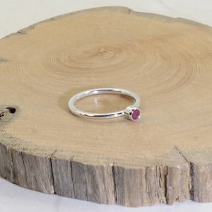 May include: A silver ring featuring a small, round, ruby-coloured gemstone. The ring is displayed on a natural wooden surface, highlighting the contrast between the polished metal and the wood's texture.