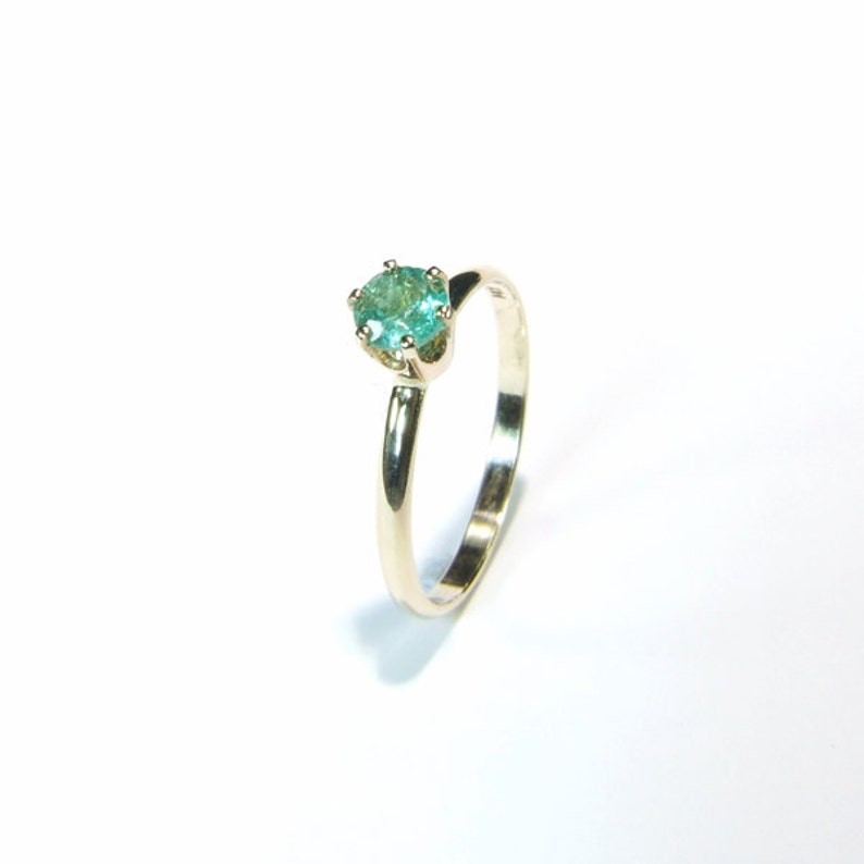 Pode incluir: Um anel de ouro com uma pedra preciosa redonda, verde claro. O anel tem uma banda fina e um design simples e elegante. A pedra preciosa &eacute; o ponto focal, definida em uma configura&ccedil;&atilde;o cl&aacute;ssica de garra.