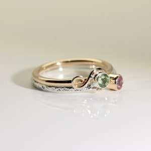 Może przedstawiać: Dwuczęściowy zestaw pierścionk&oacute;w ze złotą obrączką i srebrną obrączką. Złota obrączka ma dekoracyjny wz&oacute;r w kształcie spirali i trzyma zielony kamień szlachetny. Srebrna obrączka ma teksturowaną powierzchnię i r&oacute;żowy kamień szlachetny. Biżuteria.