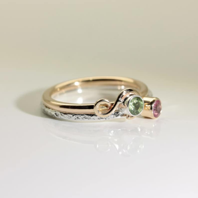 Może przedstawiać: Dwuczęściowy zestaw pierścionk&oacute;w ze złotą obrączką i srebrną obrączką. Złota obrączka ma dekoracyjny wz&oacute;r w kształcie spirali i trzyma zielony kamień szlachetny. Srebrna obrączka ma teksturowaną powierzchnię i r&oacute;żowy kamień szlachetny. Biżuteria.