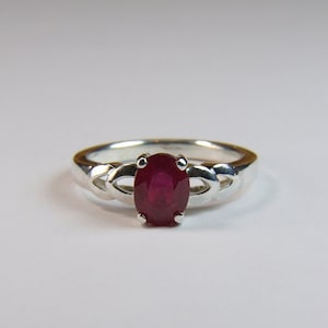 May include: A silver ring featuring a red oval ruby gemstone set in a Celtic knot design.
