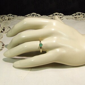 Pode incluir: Um anel de ouro com uma pedra preciosa redonda verde claro. O anel &eacute; colocado em uma fina banda de ouro. O anel &eacute; exibido em uma m&atilde;o de manequim em um fundo branco.