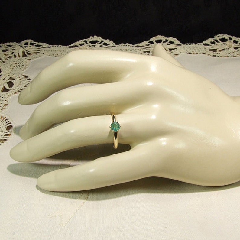 Pode incluir: Um anel de ouro com uma pedra preciosa redonda verde claro. O anel &eacute; colocado em uma fina banda de ouro. O anel &eacute; exibido em uma m&atilde;o de manequim em um fundo branco.