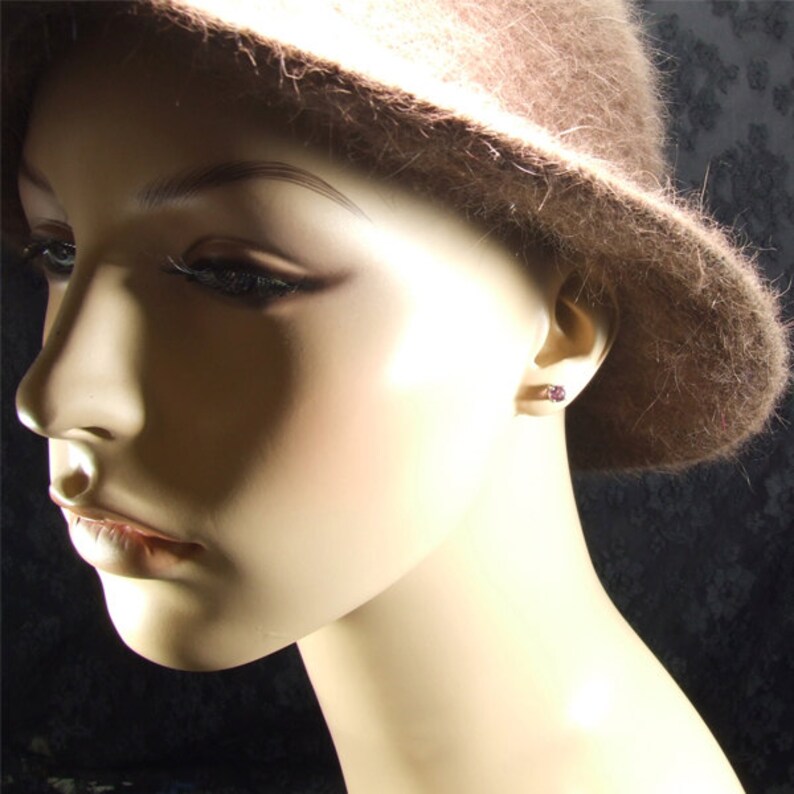 May include: A close-up of a mannequin wearing a brown felt hat and a pair of small, round, purple earrings.