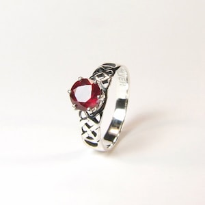 May include: A silver ring featuring a red gemstone set within a Celtic knot design. The band is inscribed with the word "SILVER".