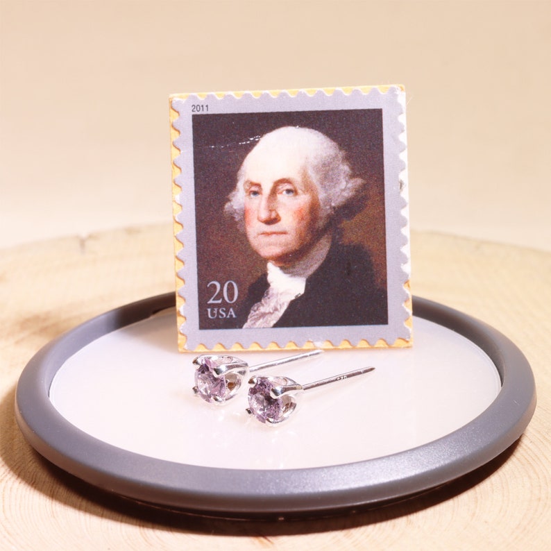 May include: A pair of silver stud earrings with small, light purple gemstones. The earrings are displayed on a white circular surface with a gray rim. A 2011 US postage stamp featuring George Washington is in the background.