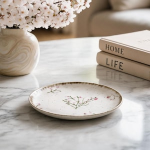 Puede incluir: Un plato de cerámica blanca con un diseño floral y un borde marrón descansa sobre una mesa de mármol. Un jarrón con flores blancas y dos libros con las palabras "HOME" y "LIFE" también están sobre la mesa.