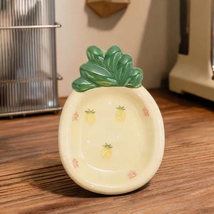 May include: A ceramic pineapple-shaped dish with a pale yellow base and green leaves. The dish features small yellow pineapple illustrations and red floral accents. The dish is on a wooden surface, with a coffee machine and utensils in the background.
