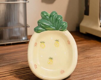 Könnte beinhalten: Eine keramische Ananas-förmige Schale mit hellgelbem Boden und grünen Blättern. Die Schale zeigt kleine gelbe Ananas-Illustrationen und rote florale Akzente. Die Schale steht auf einer Holzoberfläche, mit einer Kaffeemaschine und Utensilien im Hintergrund.