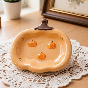 Puede incluir: Un plato de cerámica con forma de calabaza con un tallo marrón y tres detalles de calabaza naranja. El plato es de color naranja claro con lunares blancos y detalles de estrellas amarillas. Un artículo decorativo para el otoño.