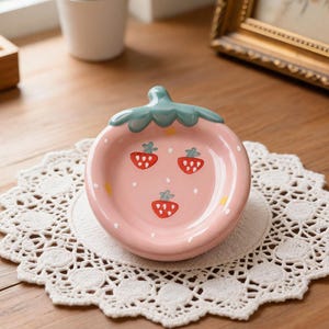 May include: A pink ceramic strawberry-shaped dish with a green stem and three red strawberry illustrations. The dish sits on a white lace doily on a wooden surface. A small potted plant and framed picture are in the background.