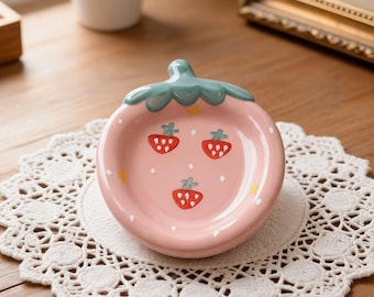Könnte beinhalten: Eine rosa Keramikschale in Erdbeerform mit einem grünen Stiel und drei roten Erdbeerillustrationen. Die Schale steht auf einer weißen Spitzendecke auf einer Holzoberfläche. Eine kleine Topfpflanze und ein gerahmtes Bild befinden sich im Hintergrund.