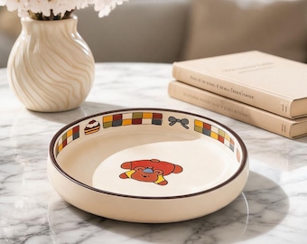 Könnte beinhalten: Eine beige Keramik-Servierschale mit braunem Rand und einer Cartoon-Bären-Illustration. Der Rand hat ein dekoratives Muster aus Quadraten, einer Schleife und einem Kuchen. Eine Vase mit weißen Blumen und zwei Bücher stehen ebenfalls auf dem Marmortisch.