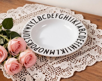 Könnte beinhalten: Ein weißer Keramikteller mit schwarzen Buchstaben am Rand liegt auf einer cremefarbenen Spitzendecke. Vier rosa Rosen sind auf einer Holzoberfläche angeordnet. Die Szene wird von weichem, natürlichem Licht beleuchtet.