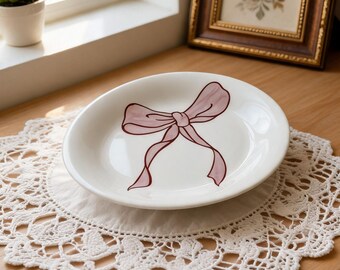 Könnte beinhalten: Ein weißer Keramikteller mit einem rosa Schleifendesign, der auf einer weißen Spitzendecke liegt. Die Decke befindet sich auf einer Holzoberfläche. Eine kleine Topfpflanze und ein gerahmtes Bild sind im Hintergrund zu sehen.