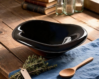 Könnte beinhalten: Eine glänzende, ovale Servierschüssel in Schwarz steht auf einem Holztisch. Ein Holzlöffel und ein Kräuterbündel mit Etikett liegen auf einem blauen Leinentuch. Bücher und Gewürzgläser sind im Hintergrund zu sehen.