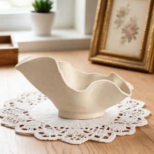 May include: A cream-colored, wavy-edged ceramic bowl sits on a white lace doily. The bowl has a unique, asymmetrical design with a flared rim. The background includes a framed picture and a small potted plant, suggesting a decorative setting.