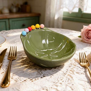 May include: A green ceramic avocado-shaped bowl with colourful round accents along the rim. The bowl sits on a lace tablecloth, with gold cutlery and a rose in the background. A unique serving dish for snacks or dips.