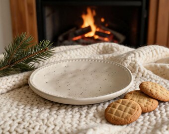Könnte beinhalten: Ein gesprenkelter, cremefarbener Keramikteller mit Perlenrand liegt auf einer cremefarbenen Strickdecke. Drei goldbraune Kekse mit Rautenmuster liegen neben dem Teller. Im Hintergrund ist ein Kamin mit brennendem Feuer zu sehen.