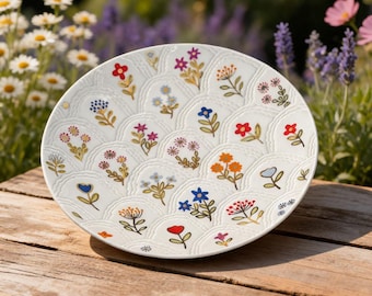 Könnte beinhalten: Ein weißer Keramikteller mit einem wellenförmigen Design und farbenfrohen Blumenillustrationen. Die Blumen sind in Rot-, Blau-, Orange- und Rosatönen gehalten. Der Teller steht auf einer Holzoberfläche, mit einem verschwommenen Blumenhintergrund.