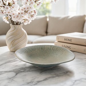 May include: A decorative, shallow, oval-shaped bowl with a textured, light green and white glaze. A vase with white and beige swirls holds cherry blossom branches. Two books with the titles "HOME" and "LIVING" are stacked on a marble table.