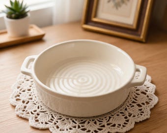 Könnte beinhalten: Eine cremefarbene, runde Keramik-Backform mit zwei Griffen, die auf einer weißen, gehäkelten Tischdecke steht. Die Form hat ein geriffeltes Innendesign. Die Szene spielt auf einer Holzoberfläche, mit einem gerahmten Bild und einer kleinen Topfpflanze im Hintergrund.