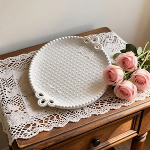 May include: A white ceramic serving plate with a textured geometric pattern and bow-shaped handles. The plate is on a wooden table with a lace doily, next to a bouquet of pink roses. The plate's diameter is approximately 25 cm.