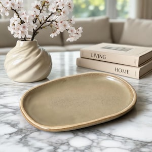 Könnte beinhalten: Eine beige ovale Servierplatte mit strukturierter Oberfläche und hellbraunem Rand. Eine weiße Vase mit Spiralmuster hält Kirschblütenzweige. Zwei Bücher mit den Worten "LIVING" und "HOME" sind im Hintergrund gestapelt.