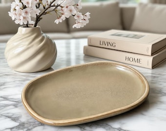 May include: A beige oval serving platter with a textured surface and a light brown rim. A white vase with a spiral design holds cherry blossom branches. Two books with the words "LIVING" and "HOME" are stacked in the background.