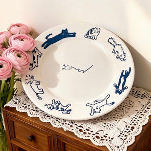 May include: White ceramic plate decorated with navy blue cat illustrations. The plate showcases various cat poses around the rim, creating a whimsical and artistic aesthetic. A charming addition to any kitchen or dining setting.