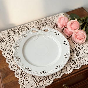 May include: A white ceramic plate featuring a bow design and floral cutouts around the rim. The plate sits on a lace doily, with pink roses nearby. Ideal for serving food or as a decorative item. The plate's design adds a touch of elegance.
