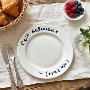 May include: A white ceramic plate with the words "C'est délicieux ~ (tres bon)" in blue script. The plate is on a white linen tablecloth, with a basket of croissants, a small bowl of berries, and silverware.