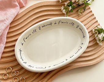 Könnte beinhalten: Eine cremefarbene ovale Servierplatte aus Keramik mit schwarzer Schrift am Rand, die "à Paris est il minuit" lautet. Die Platte liegt auf einem Holztablett mit gewelltem Rand, mit goldenen Ringen und Blumen.