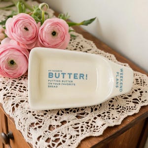 May include: A cream-coloured rectangular butter dish with blue text reading "MY FAVORITE BUTTER! PUTTING BUTTER ON YOUR FAVORITE BREAD". The words "WEEKEND PLANS" are printed on the side. Pink flowers are in the background.