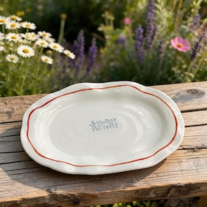 Puede incluir: Plato de cerámica blanco con borde ondulado y borde rojo. El plato tiene el texto "SUNDAY POTTERY" en escritura azul. El plato está sobre una superficie de madera, con flores en el fondo.